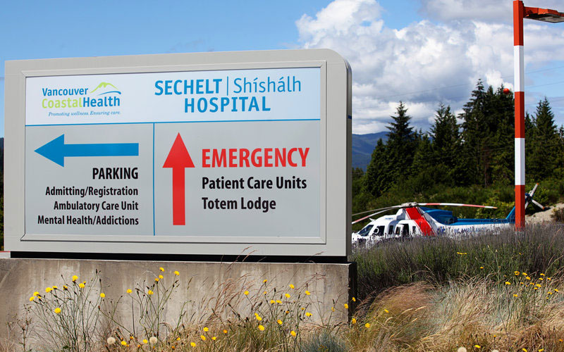 Facility-Upgrades-final Vancouver Coastal Health/ Sechelt | Shíshálh Hospital sign with directional arrows labeled “Parking” and “Emergency,” with a BC Ambulance helijet visible in the background.