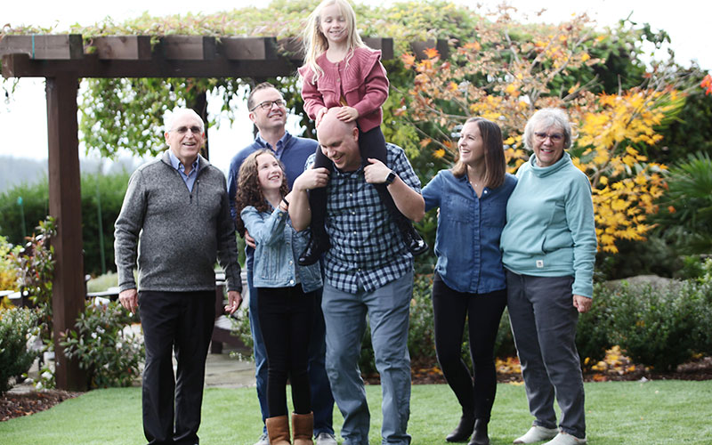 Generation2Generation-final A multi-generational family smiling outdoors during a family photo, with the youngest girl sitting on her dad’s shoulders.