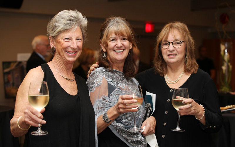 Host-A-Fundraiser-final Three women dressed in evening attire smile and hold glasses of white wine at a social fundraising event.