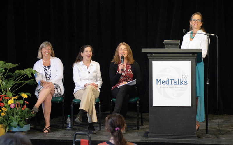 MedTalks-communities Four women participate in a MedTalks health and wellness panel discussion; three are seated and one stands at a podium speaking to the audience in front of a black backdrop with flowers on stage.