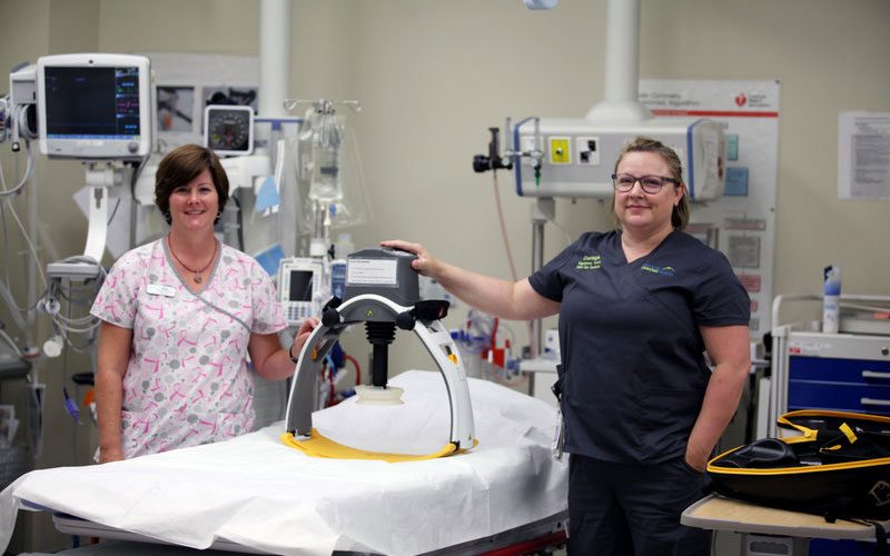 News-final Two healthcare professionals in a hospital room demonstrate a mechanical CPR device positioned on a patient bed, surrounded by medical monitors and equipment.