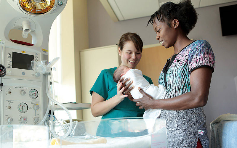 Vital-Equipment-final nurse hands a newborn baby to another smiling nurse in a hospital room beside an infant warmer and medical equipment.